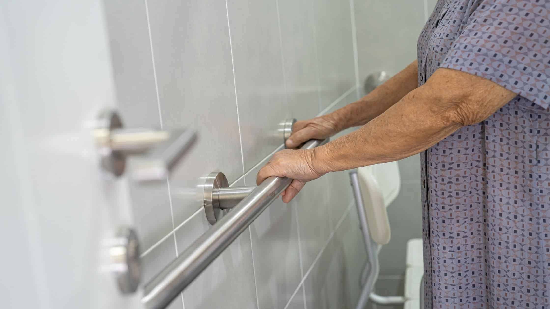 Elderly person holding a stainless steel grab bar in a bathroom for safety and support.